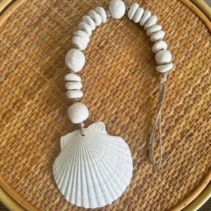 May include: White seashell garland with a large white seashell hanging from the bottom. The garland is made of white wooden beads and a natural twine string.