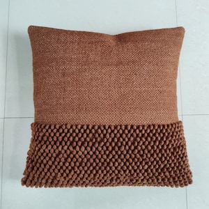 May include: A brown throw pillow with a textured fabric top and a brown pom-pom trim around the bottom edge.