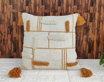 May include: A white throw pillow with a geometric pattern in mustard yellow. The pillow has fringe tassels on each corner.