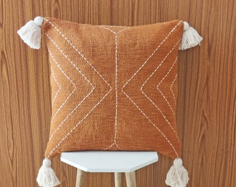 Puede incluir: Una almohada de tiro de color naranja quemado con costuras geométricas blancas y borlas blancas en cada esquina. La almohada está sentada en un taburete blanco con patas de madera.