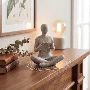 Pode incluir: Uma estatueta de pedra cinzenta salpicada de uma pessoa sentada de pernas cruzadas enquanto segura um livro aberto. A escultura mede 16,5 cm de altura, 13,5 cm de largura e 8,5 cm de profundidade, conforme indicado pelas linhas de medição.