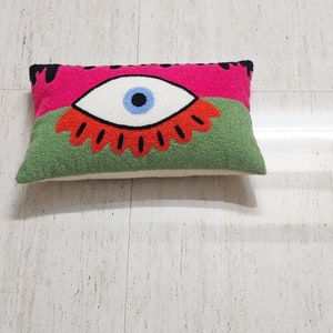 May include: A green and pink pillow with an embroidered eye design. The eye is blue and white with a red and orange flower around it.