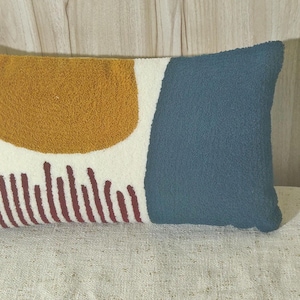 Puede incluir: Cojín decorativo rectangular con superficie texturizada. Presenta un semicírculo amarillo mostaza, un rectángulo azul verdoso y líneas verticales burdeos sobre fondo blanco. Para decoración del hogar.
