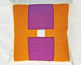 Puede incluir: Una almohada blanca con un diseño geométrico que presenta una gran forma de "H" púrpura sobre un fondo naranja brillante.