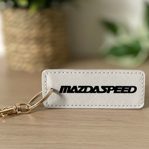 Puede incluir: Llavero de cuero blanco con un llavero dorado y el texto "MAZDASPEED" en letras negras.