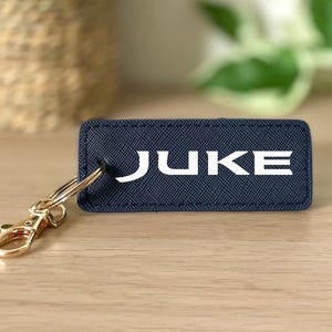 Puede incluir: Un llavero azul marino con un anillo de metal dorado y un clip. El llavero tiene una etiqueta de tela blanca con la palabra "JUKE" impresa en ella.