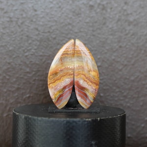 Pode incluir: Um par de cabochões em forma de lágrima com um padrão em camadas de marrom, laranja e branco. Os cabochões são polidos e têm um acabamento brilhante.