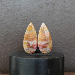 May include: Two teardrop-shaped gemstones with a polished finish. The stones display a layered pattern of warm colors, including yellow, orange, red, and white. The gemstones are displayed on a black cylindrical stand against a neutral background.