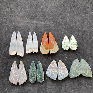 May include: Assortment of gemstone cabochons in various shapes and colors, including teardrop and triangular forms. The stones feature patterns in shades of white, green, orange, and brown. The cabochons are displayed on a dark gray surface.