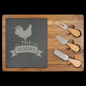 Puede incluir: Una tabla de servir rectangular de madera con una incrustación de pizarra gris oscuro que presenta un gráfico de gallo grabado y el texto "Est. 1928 THE OLSON'S". Tres cuchillos de queso de acero inoxidable con mangos de madera clara.