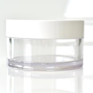 Op de afbeelding: Een transparante plastic pot met een wit deksel. De pot is leeg en heeft een gladde, ronde vorm.