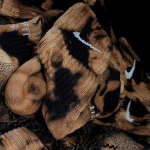 Puede incluir: Primer plano de un montón de calcetines de color marrón y negro con efecto tie-dye y logotipos Nike blancos. Los calcetines están dispuestos de forma aleatoria y están rodeados de papel triturado negro.