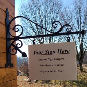 May include: Black wrought iron sign holder with decorative scrollwork, mounted on a wooden wall. A white rectangular sign is suspended from the holder, with the text "Your Sign Here". The sign offers custom sign hangers, with designs up to 3 feet by 4 feet.