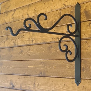 May include: Black wrought iron hanging bracket with decorative scrollwork. The bracket is mounted on a wooden wall with horizontal planks. The bracket has a vertical mounting plate and a horizontal arm with a curved end.