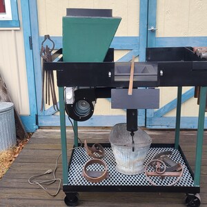 May include: A metal and wood workshop cart with a green hopper, black table, and a metal bucket. The cart has a metal grate shelf and is on wheels. Tools and decorative metal pieces are on the shelf.
