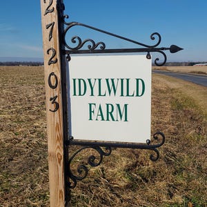 May include: A wooden post with the address 27203 and a sign that reads "IDYLWILD FARM" in green lettering. The sign is white with black decorative metalwork and is set against a blue sky.