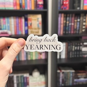 以下が含まれることがあります： 黒い筆記体で「bring back yearning」と書かれた白いステッカー。ステッカーは本でいっぱいの本棚の前に置かれています。