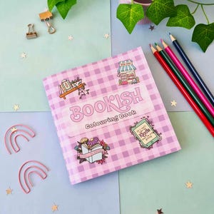 May include: A "Bookish Colouring Book" with a pink gingham cover, decorated with illustrations of a bookstore, flowers, and a Kindle. The image also includes colored pencils, paper clips, and decorative stars.