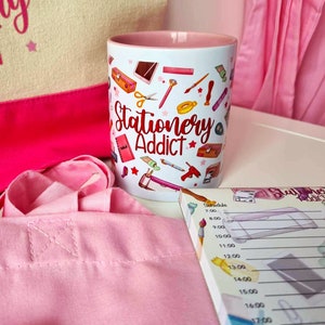 May include: A pink and white mug with the text "Stationery Addict" and a pattern of various stationery items. The mug is sitting on a white surface next to a pink tote bag and a pink and white notepad with a similar stationery pattern.