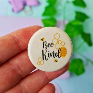 May include: White button with a yellow and black bee design. The button says "Bee Kind" with a honeycomb pattern.