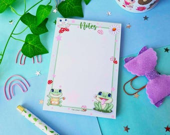 May include: A notepad with a green, pink, and white border featuring a frog illustration and the word "Notes". The notepad is on a blue surface with a purple bow and a green plant.