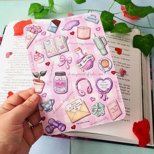 May include: A pink and white sticker sheet featuring illustrations of a cupcake, a jar labelled "Self-Love", and the phrases "I am enough" and "Love Yourself First". The sheet is held over an open book.