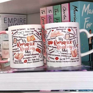 May include: Two white ceramic mugs with pink handles. Both mugs feature a pink and red design with the text "My Romance Reading Mug" and various romance novel tropes. The mugs are on a shelf with books behind them.