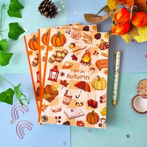 May include: Three notebooks with orange covers decorated with autumn-themed illustrations, including pumpkins and leaves. The text "I am an Autumn" is printed on the cover. A pen and decorative tape rolls are also visible.
