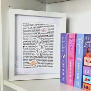 May include: A white framed print with a list of romance novel tropes in black text with pink hearts. The print is sitting on a shelf with three books titled "Icebreaker", "Wildfire", and "Daydream" by Hannah Grace. The books are stacked on top of two other books titled "Daydream" and "Good" by Elle Kennedy.
