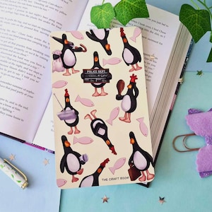 May include: A cream-colored notebook featuring cartoon penguins in various poses, with pink fish accents. The notebook has the text "POLICE DEPT" and "THE CRAFT ROOM". The notebook is open on a book.
