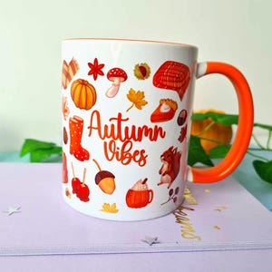 May include: White ceramic mug with an orange handle and rim, decorated with autumn-themed illustrations. The mug features images of pumpkins, leaves, and the text "Autumn Vibes". The mug is on a purple surface.