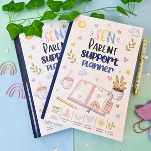 May include: Two white planners titled SEN Parent Support Planner with dark blue spines. The covers feature colorful illustrations of a sun, clouds, hearts, stars, a coffee mug, a potted plant, and an open notebook with a pencil and rainbow icon.