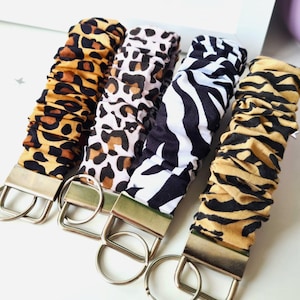May include: Four fabric keychains with animal print patterns: leopard, cheetah, zebra, and tiger. Each keychain has a silver-colored metal clasp and ring. The fabric is gathered, creating a textured appearance.