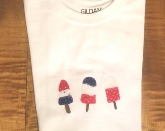 May include: White t-shirt with a red, white, and blue popsicle design. The shirt has the word "Gildan" printed on the neck.
