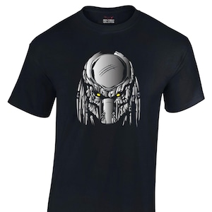 Puede incluir: Camiseta negra con una ilustración detallada en plata y gris de la cara de un Depredador. El diseño incluye ojos amarillos y detalles faciales intrincados. La camiseta es 100% algodón y tiene la marca "Red Chilli" impresa.