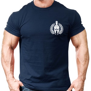 以下が含まれることがあります： 紺色のTシャツに、月桂樹の輪の中にスパルタのヘルメットが描かれた白いグラフィックがプリントされています