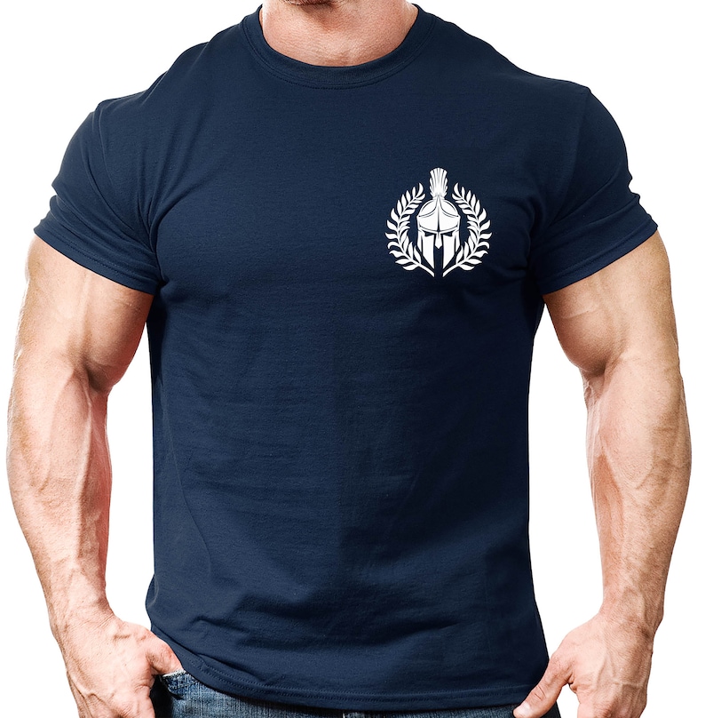 以下が含まれることがあります： 紺色のTシャツに、月桂樹の輪の中にスパルタのヘルメットが描かれた白いグラフィックがプリントされています