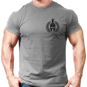 以下が含まれることがあります： 左胸に黒いスパルタのヘルメットのグラフィックが描かれたグレーのTシャツ。ヘルメットは月桂樹の輪で囲まれています。