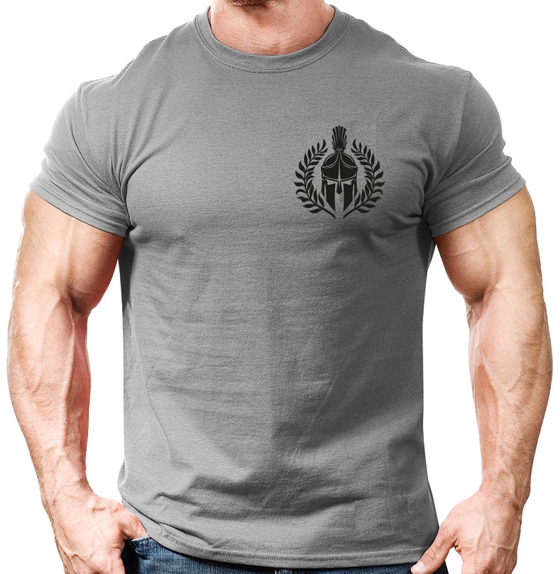以下が含まれることがあります： 左胸に黒いスパルタのヘルメットのグラフィックが描かれたグレーのTシャツ。ヘルメットは月桂樹の輪で囲まれています。