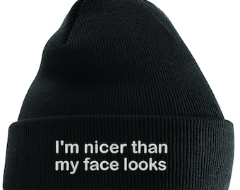 May include: Black beanie hat with the embroidered text "I'm nicer than my face looks" in white. The hat is made of a knitted material and has a folded brim. A casual accessory for cold weather.