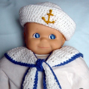 May include: A white crocheted sailor hat with a yellow anchor, a white crocheted sailor collar with blue trim, and a white crocheted scarf with blue trim. The hat and scarf are worn by a doll.