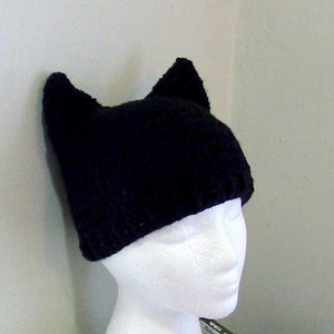 K&ouml;nnte beinhalten: Schwarze H&auml;kelm&uuml;tze mit Katzenohren. Die M&uuml;tze ist aus weichem, warmem Garn gefertigt und perfekt, um Kopf und Ohren im Winter warm zu halten.