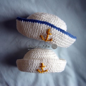 May include: Two white crocheted sailor hats with blue trim and yellow anchor embroidery. The hats are different sizes, one is larger than the other.