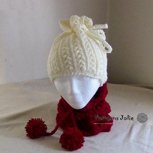 Op de afbeelding: Een witte gehaakte muts met een grote strik bovenop en een rode gehaakte nekwarmer met twee pompoenen. De tekst "Patterns Jolie" is zichtbaar op de nekwarmer.