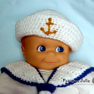 May include: A white crocheted sailor hat with a gold anchor on the front, worn by a doll with blue eyes. The doll is wearing a white crocheted sailor collar with a blue trim.