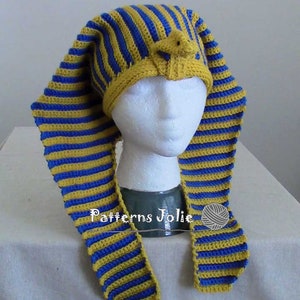 K&ouml;nnte beinhalten: Eine gestrickte M&uuml;tze mit gelben und blauen Streifen und einer kleinen, gelben, geh&auml;kelten Figur auf der Oberseite. Die M&uuml;tze hat lange, flie&szlig;ende Streifen, die am Nacken herunterh&auml;ngen. Die M&uuml;tze sitzt auf einem wei&szlig;en Mannequin-Kopf.