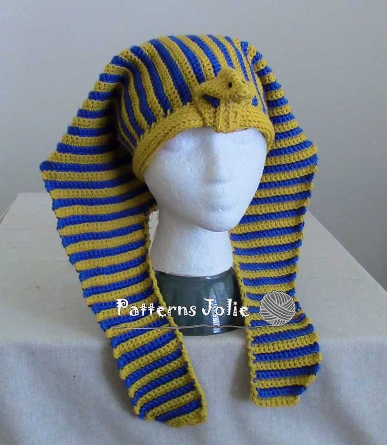 K&ouml;nnte beinhalten: Eine gestrickte M&uuml;tze mit gelben und blauen Streifen und einer kleinen, gelben, geh&auml;kelten Figur auf der Oberseite. Die M&uuml;tze hat lange, flie&szlig;ende Streifen, die am Nacken herunterh&auml;ngen. Die M&uuml;tze sitzt auf einem wei&szlig;en Mannequin-Kopf.