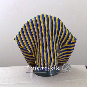 K&ouml;nnte beinhalten: Eine geh&auml;kelte M&uuml;tze mit blauen und gelben Streifen. Die M&uuml;tze befindet sich auf einem Pr&auml;sentationsst&auml;nder. Der Text "Patterns Jolie" ist unter der M&uuml;tze sichtbar.