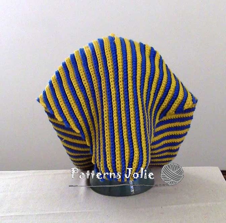K&ouml;nnte beinhalten: Eine geh&auml;kelte M&uuml;tze mit blauen und gelben Streifen. Die M&uuml;tze befindet sich auf einem Pr&auml;sentationsst&auml;nder. Der Text "Patterns Jolie" ist unter der M&uuml;tze sichtbar.
