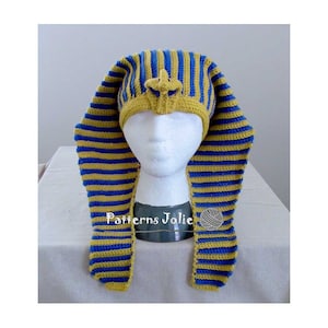 Könnte beinhalten: Eine gehäkelte Mütze mit einem blau-gelben Streifenmuster, die an die Kopfbedeckung eines ägyptischen Pharaos erinnert. Die Mütze wird auf einem weißen Mannequin-Kopf präsentiert. Der Text "Patterns Jolie" ist auf dem Sockel des Mannequins sichtbar.